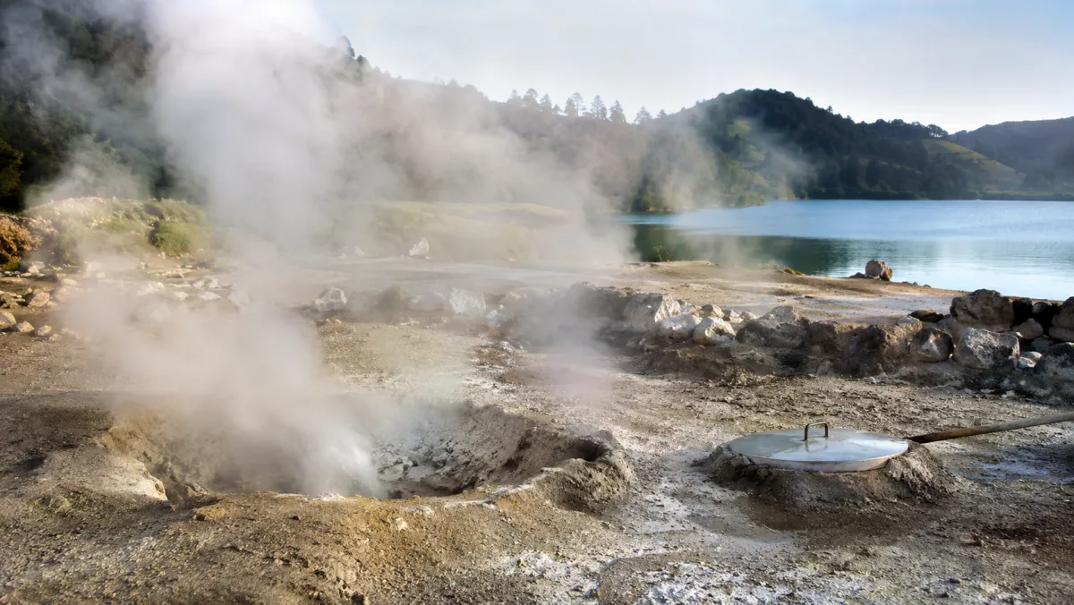 Fumarolas humeantes junto a la Lagoa das Furnas con una olla de cozido enterrada en el suelo geotérmico ocre y la tapa metálica asomando, São Miguel, Azores