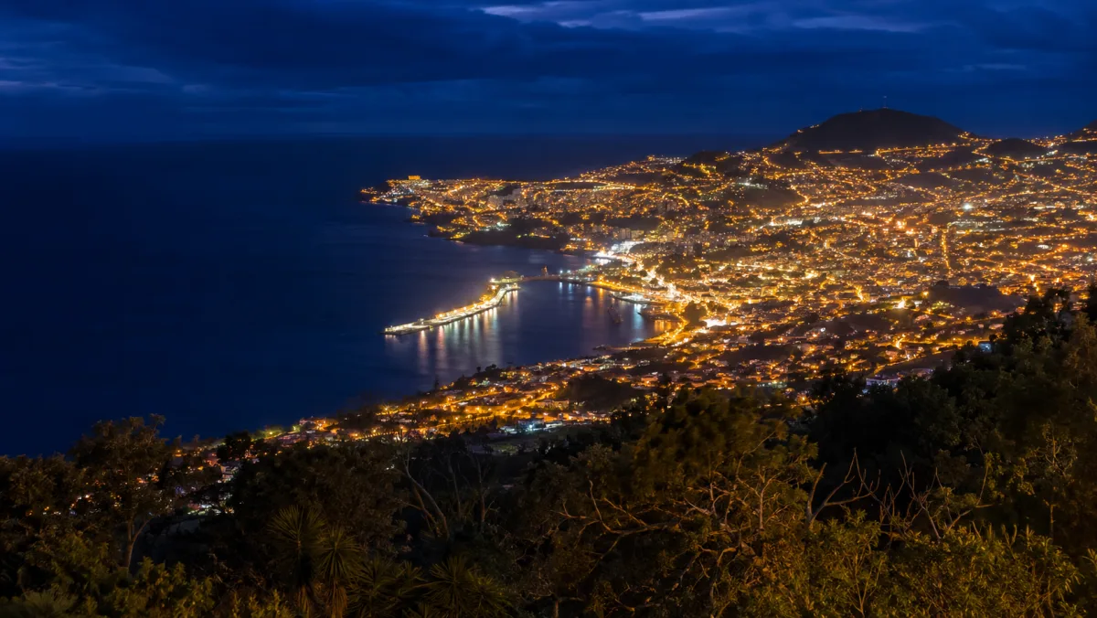 Vista nocturna desde las alturas sobre Funchal: un manto de luces doradas curvándose alrededor de la bahía oscura, con el puerto iluminado reflejándose en el agua