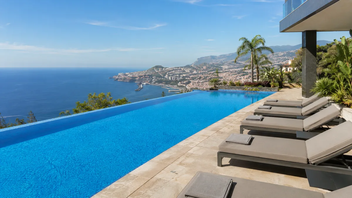 Terraza de hotel con piscina infinita de borde desbordante sobre la bahía de Funchal, tumbonas, palmeras y la ciudad y el Atlántico al fondo en un día despejado