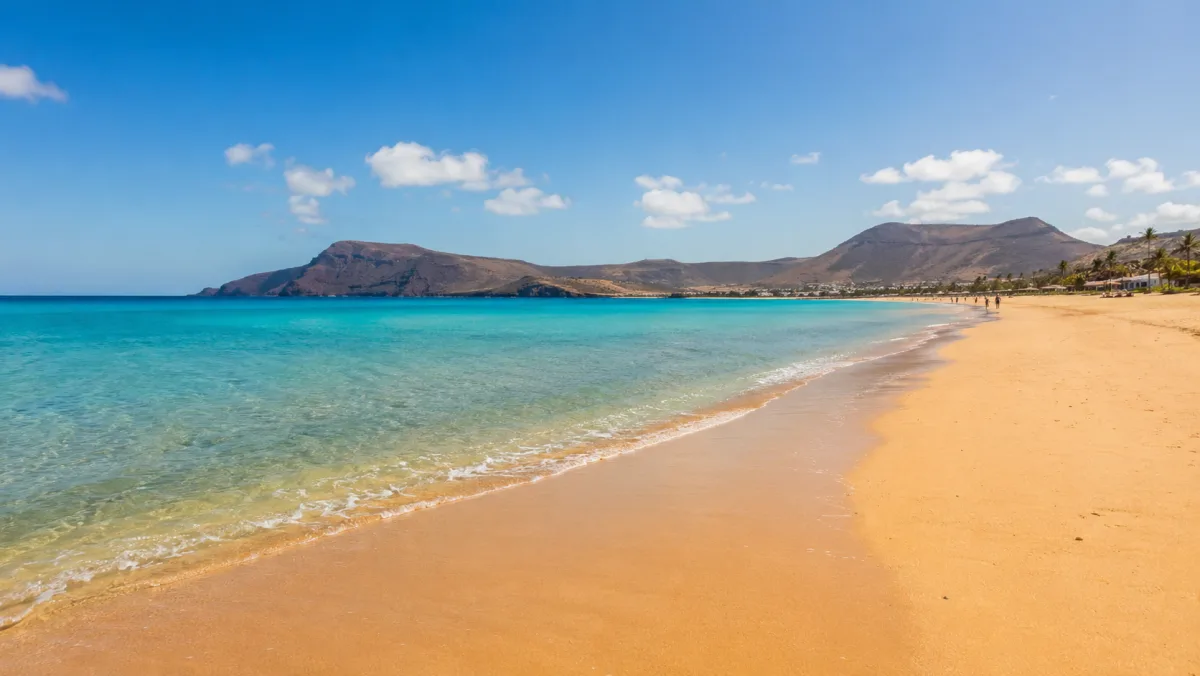 La larga playa de arena dorada de Porto Santo con agua turquesa transparente y las montañas de la isla al fondo bajo un cielo despejado