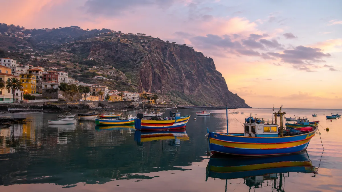 El puerto de Câmara de Lobos al amanecer con barcas de pesca de colores reflejadas en el agua en calma y un gran acantilado al fondo
