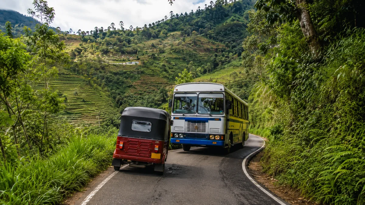 Un tuk-tuk rojo y un bus cruzándose en una estrecha carretera de montaña con curvas rodeada de vegetación verde, camino de Ella, Sri Lanka