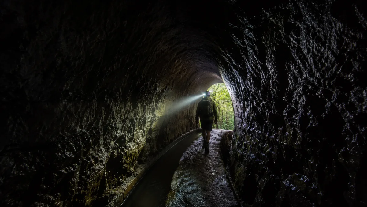 Interior oscuro de un túnel de la Levada do Caldeirão Verde: un senderista con linterna frontal ilumina las paredes de roca húmeda y al fondo se ve el bosque verde