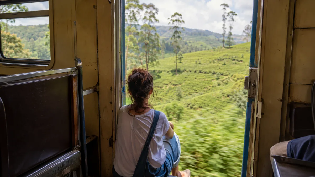 Una viajera sentada de espaldas en el marco de la puerta abierta de un vagón del tren de Kandy a Ella, mirando pasar las colinas verdes de plantaciones de té, Sri Lanka