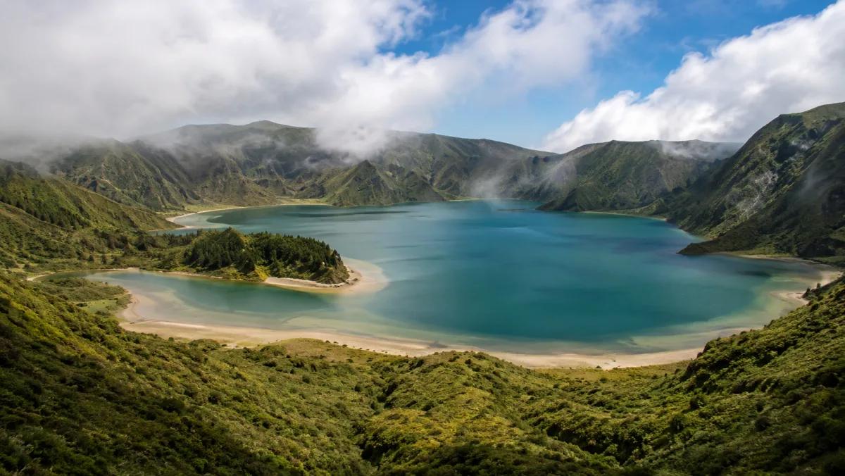 La Lagoa do Fogo, lago de cráter de aguas verde-azuladas con orilla de arena clara, rodeado de laderas verdes empinadas y jirones de niebla, en el corazón de São Miguel, Azores