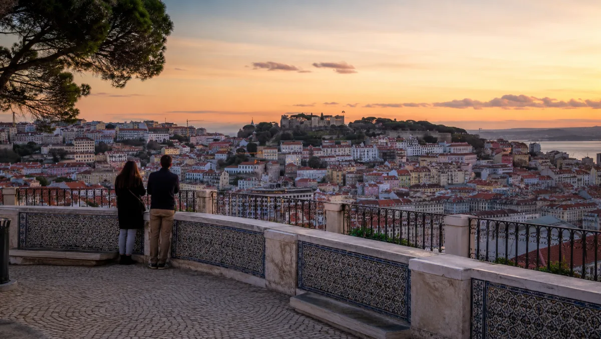 Una pareja de espaldas apoyada en la balaustrada de azulejos de un mirador contempla el casco antiguo de Lisboa y el castillo al atardecer