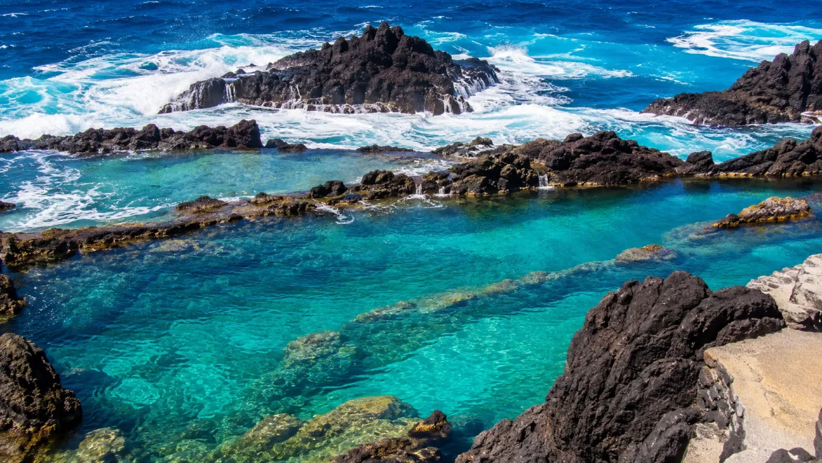 Piscinas naturales de lava de Porto Moniz con agua turquesa transparente entre roca volcánica negra y el Atlántico rompiendo en blanco detrás