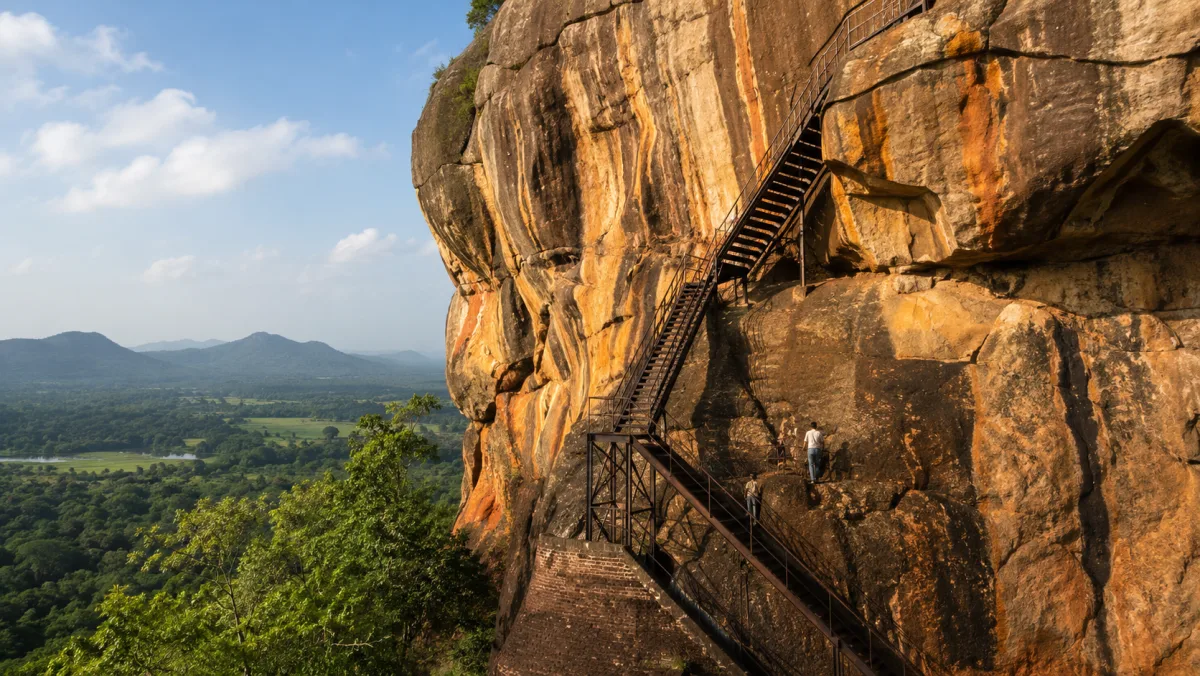 Pared de roca de la fortaleza de Sigiriya con las escaleras metálicas subiendo entre la piedra ocre y la llanura verde al fondo, Sri Lanka
