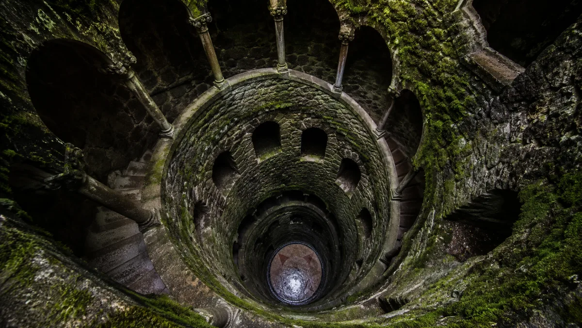 El pozo iniciático de la Quinta da Regaleira visto desde arriba: una escalera de caracol de piedra cubierta de musgo desciende en espiral hacia el fondo