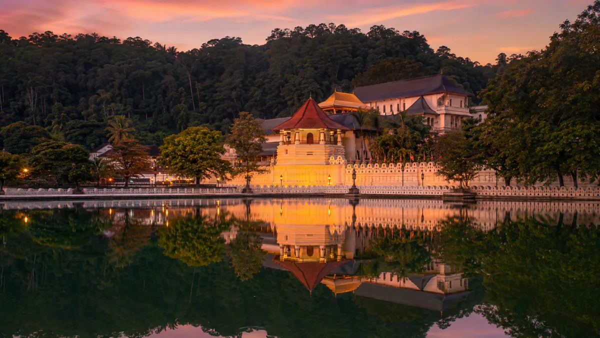 El Templo del Diente (Sri Dalada Maligawa) iluminado al atardecer y reflejado en el lago de Kandy, con las colinas verdes de fondo, Sri Lanka